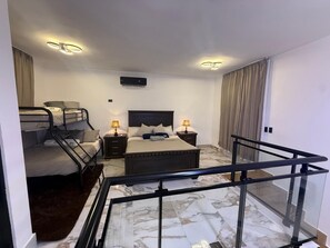Quarto de hotel com cama, secretária, cadeira, janela com cortinas, luz no teto e escada com grade de vidro.