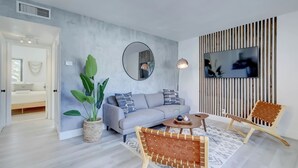 Ein modernes Wohnzimmer mit einem grauen Sofa, einem runden Couchtisch, Korbstühlen, einem großen Spiegel und einem Fernseher, der an einer Holzwand befestigt ist.