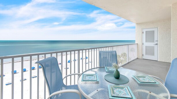 Ein Balkon mit Blick auf Strand und Meer, ein runder Glastisch mit Stühlen und eine Tür mit Fensterläden.