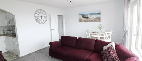 A living room with a red sofa, a dining table with chairs, a kitchen area, and a large window.