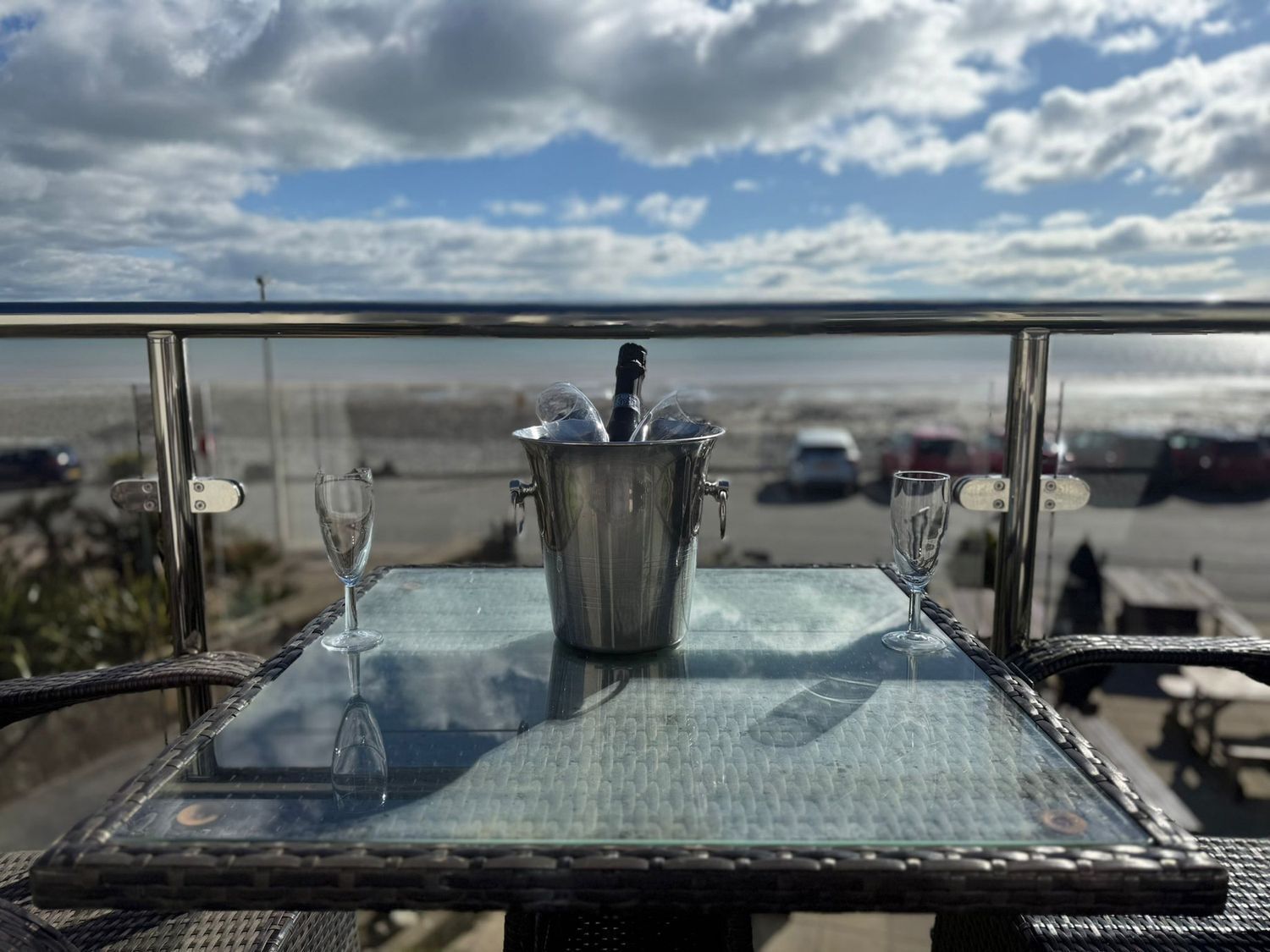 Una mesa con un balde metálico que contiene una botella y vasos, con vistas a una playa con coches aparcados y un cielo nublado.