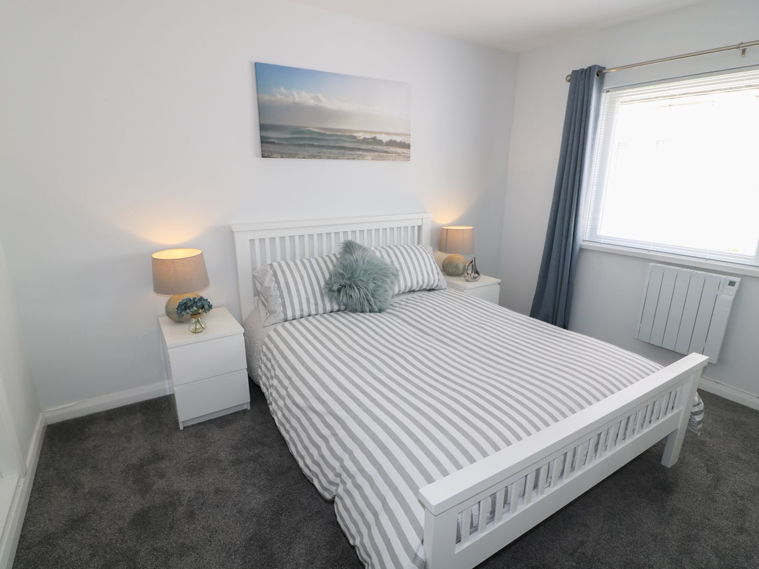 A bedroom with a bed, two bedside tables, a nightstand, a window with curtains, and a wall-mounted picture.