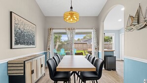 Un espace repas avec une longue table en bois, des chaises noires, un luminaire suspendu et une vue sur l’extérieur, accessible par des portes vitrées.