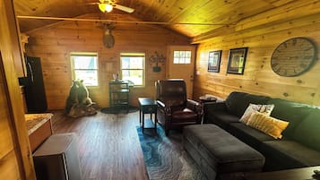 A cozy cabin interior with a wooden ceiling, a ceiling fan, a dining table, a leather armchair, a sofa, and a clock on the wall.