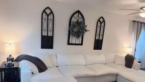 Un salon moderne avec un canapé d’angle, deux fenêtres en arc fixées au mur et un ventilateur de plafond.