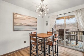 A dining area with a wooden table and chairs, a large abstract painting, a ceiling light fixture, and a sliding glass door leading to an outdoor patio. - Lake Taneycomo View -  Cozy 1BR Condo - Fireplace (Branson)