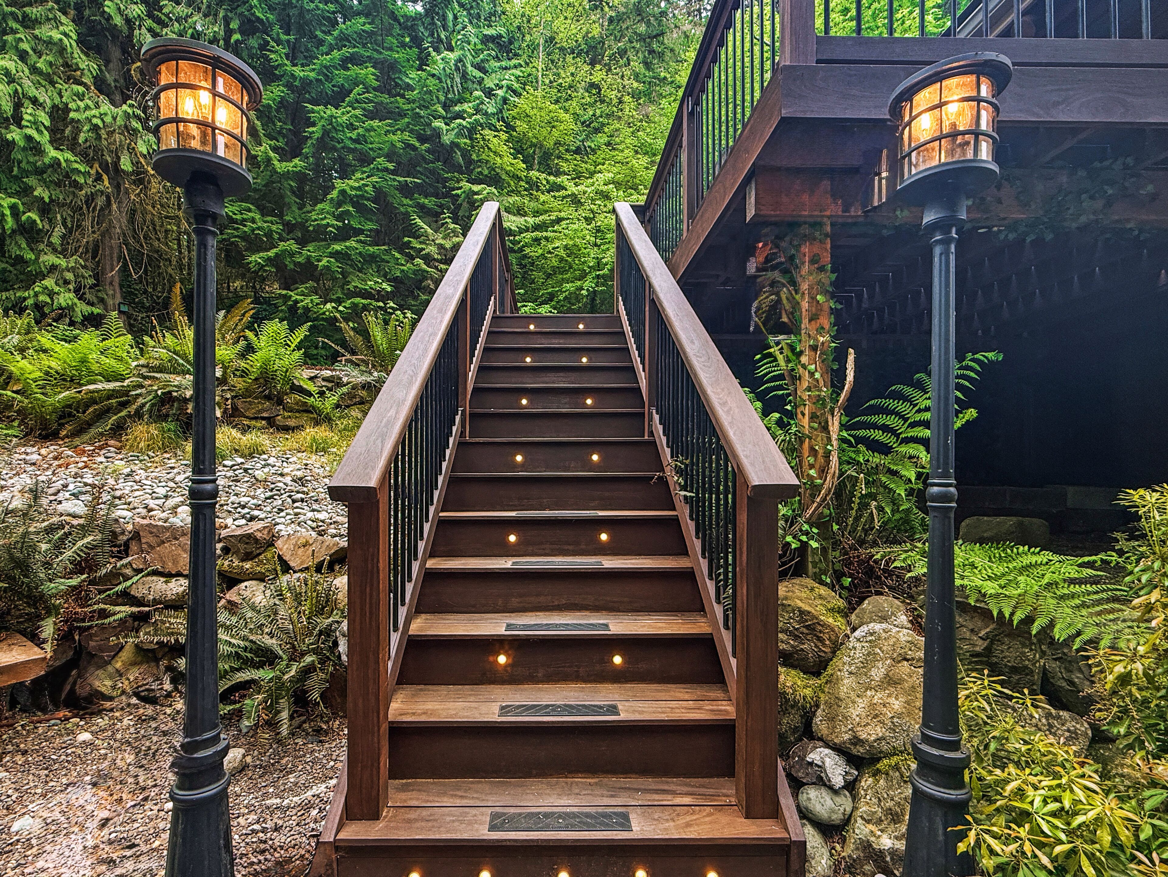 A wooden staircase with railings and lights, surrounded by greenery and rocks.