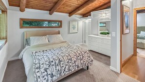 Een slaapkamer met een houten plafond, een bed met beddengoed met een patroon, een dressoir en een raam met uitzicht op groen.