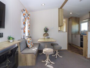 A modern kitchen with a built-in oven, a dining area with a sofa and stools, and a small table with a vase of flowers. - 213 Seaview Caravan (Paignton)