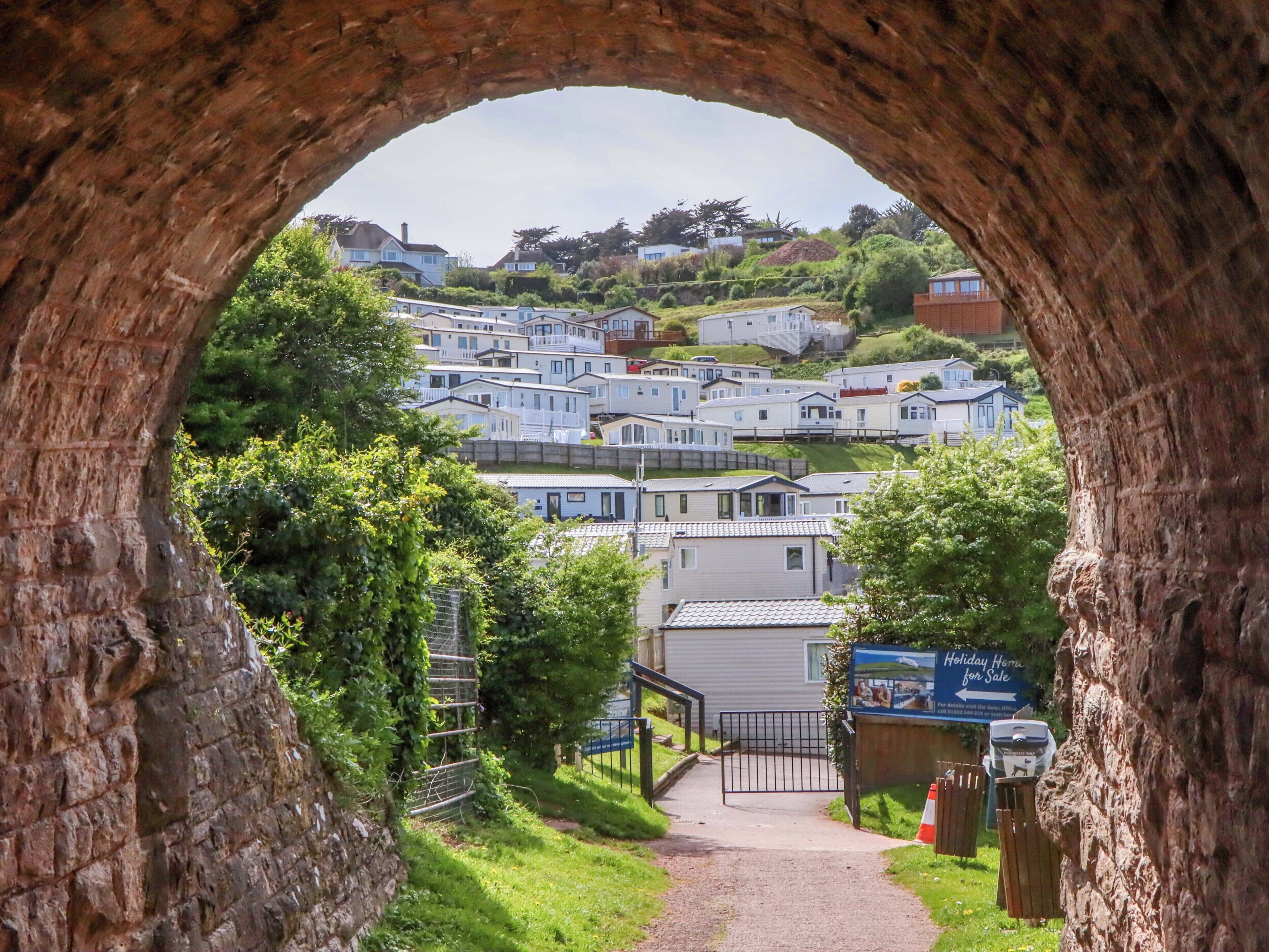 A view through a stone archway of a residential area with multiple houses and a sign for a holiday home for sale.