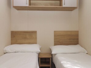 A room with two beds, each with a wooden headboard, a small wooden nightstand, and a white cabinet above the beds. - 213 Seaview Caravan (Paignton)