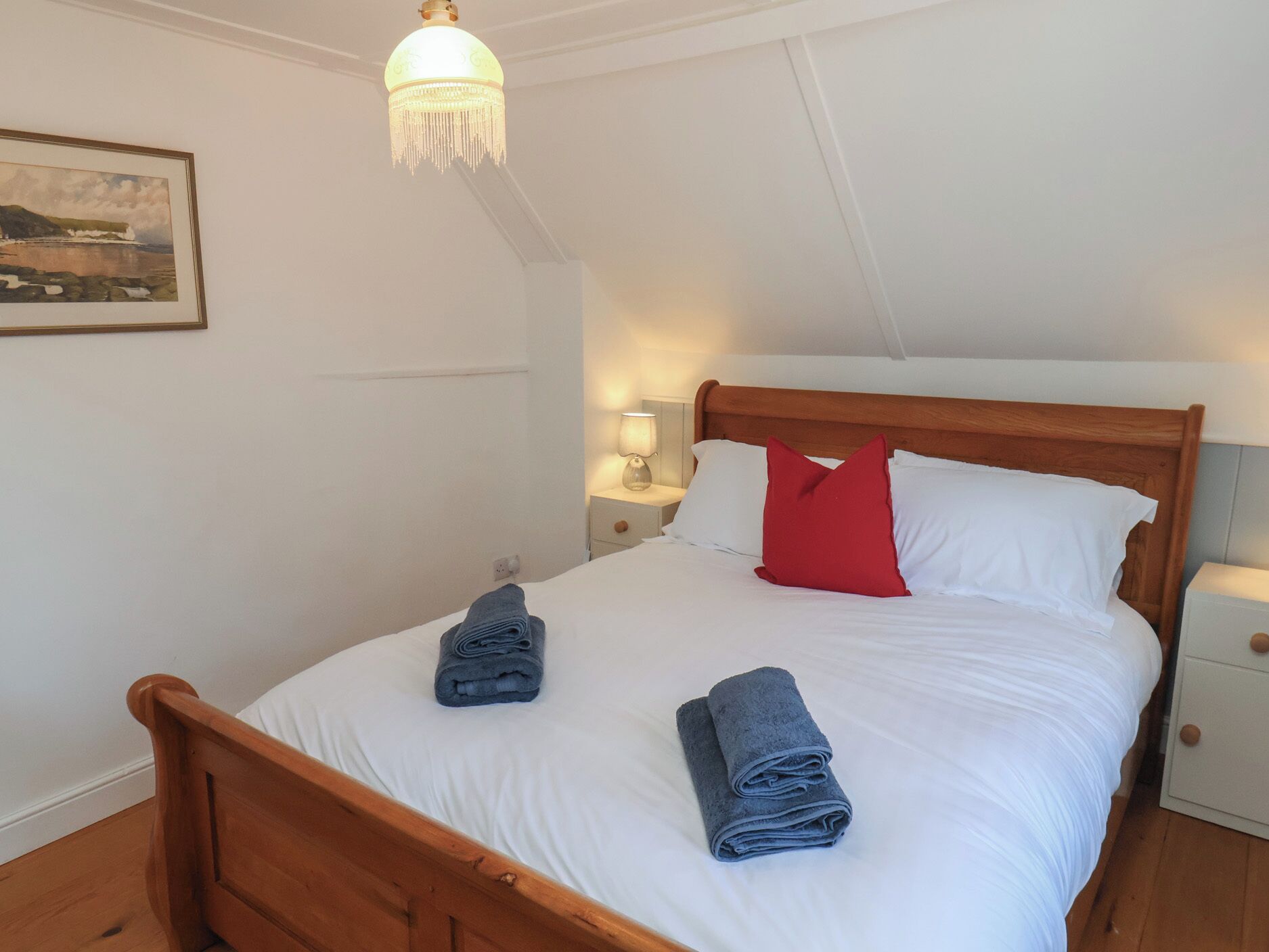 Ein Schlafzimmer mit einem hölzernen Bett, weißer Bettwäsche, einem roten Kopfkissen und zwei gerollten Handtüchern.