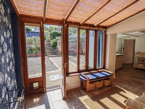 A room with a sliding glass door leading to a garden, a wooden bench with storage baskets, and a kitchen area visible through the door. - Fleming Cottage (Filey)