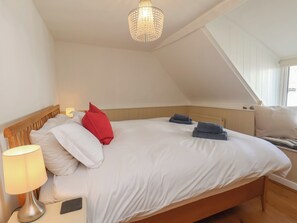 A bedroom with a wooden headboard, white bedding, a red pillow, a lamp, and a chandelier. - Fleming Cottage (Filey)