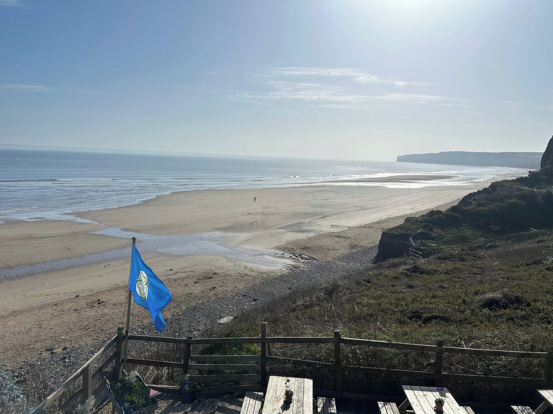 Blick auf die Küste mit einem Sandstrand, einem Holzzaun und einer Fahne.