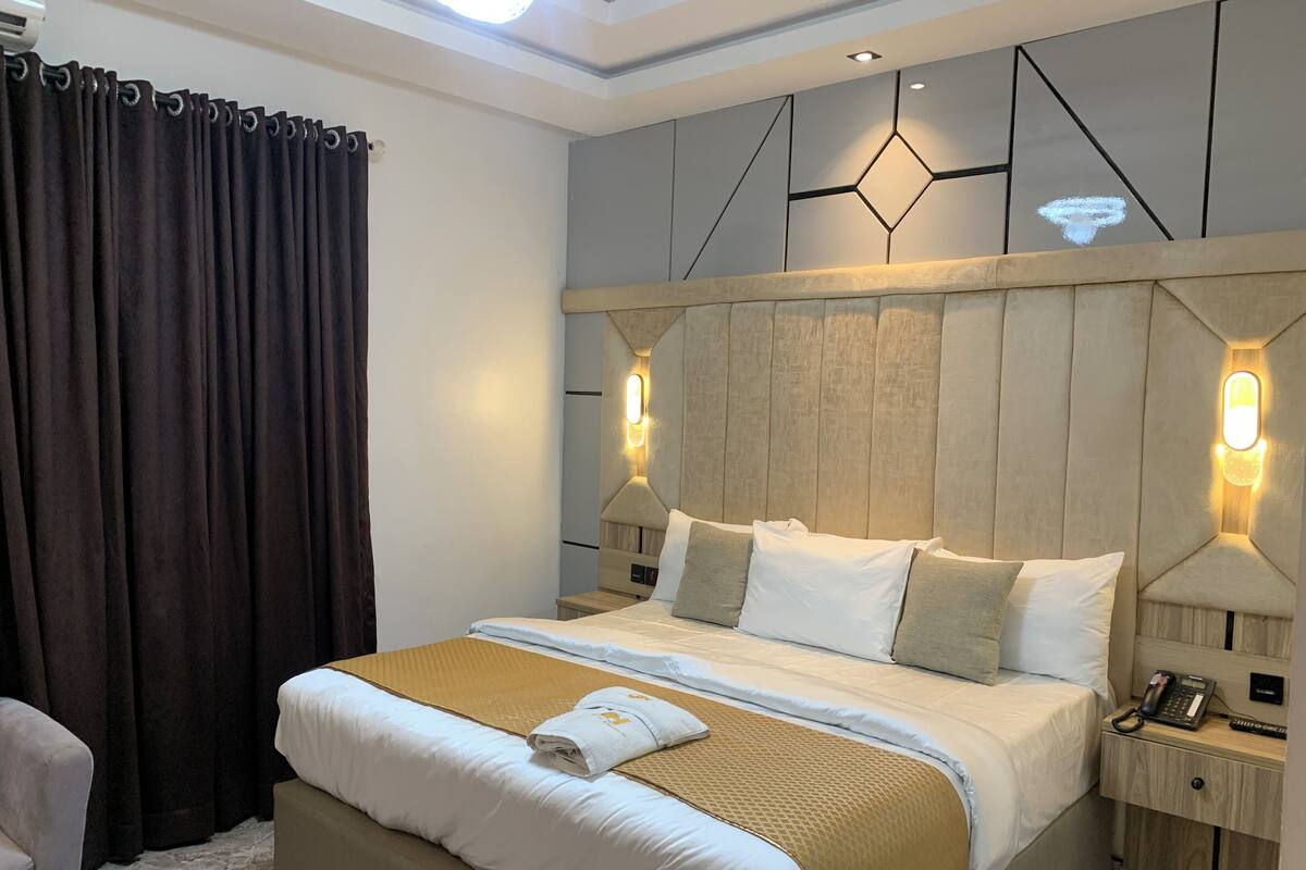 Una habitación de hotel moderna con una cama grande, una mesita de noche con teléfono y un cabecero con paneles geométricos en la pared.