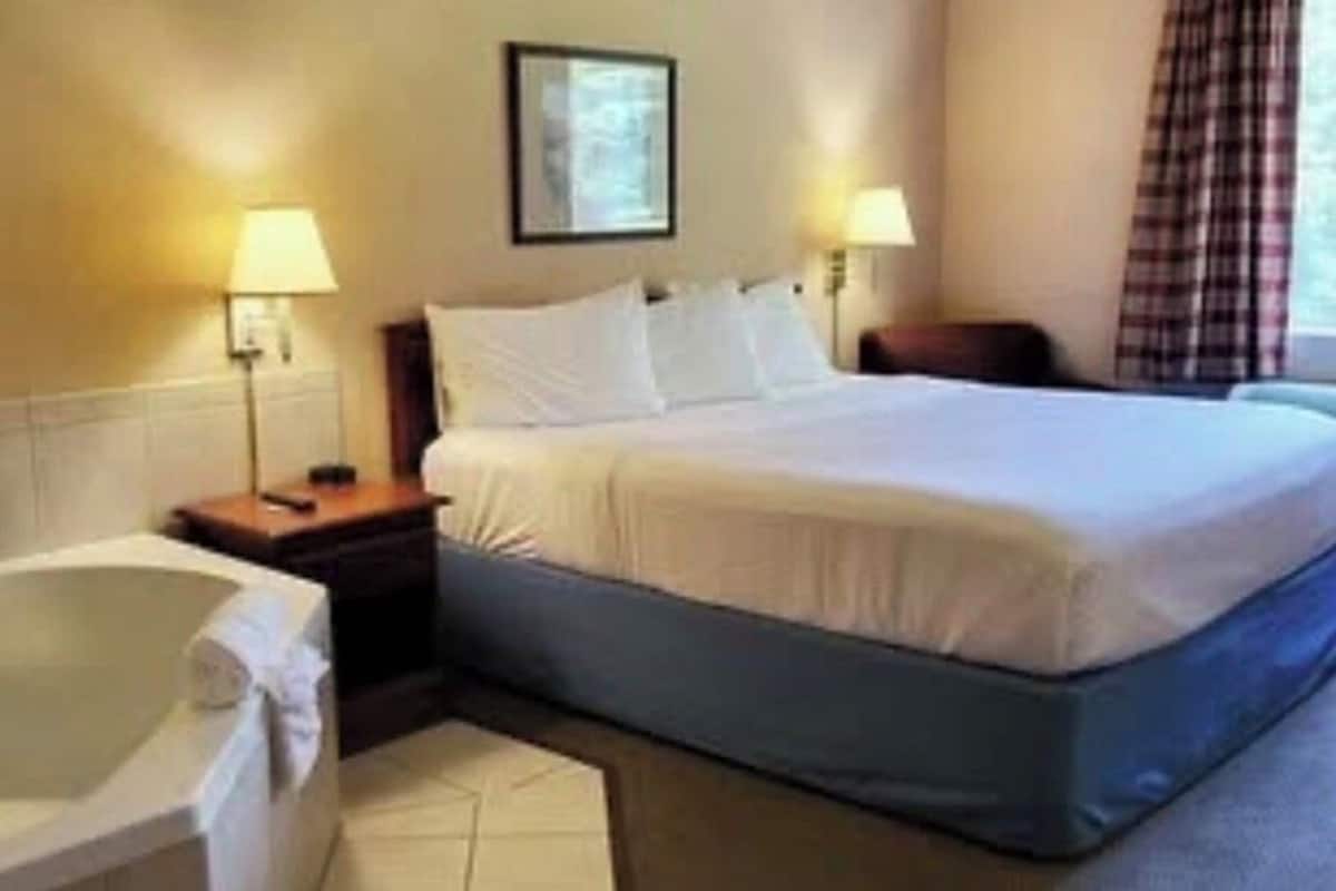 Quarto de hotel com cama, mesa de cabeceira, abajur, janela com cortinas e banheira.