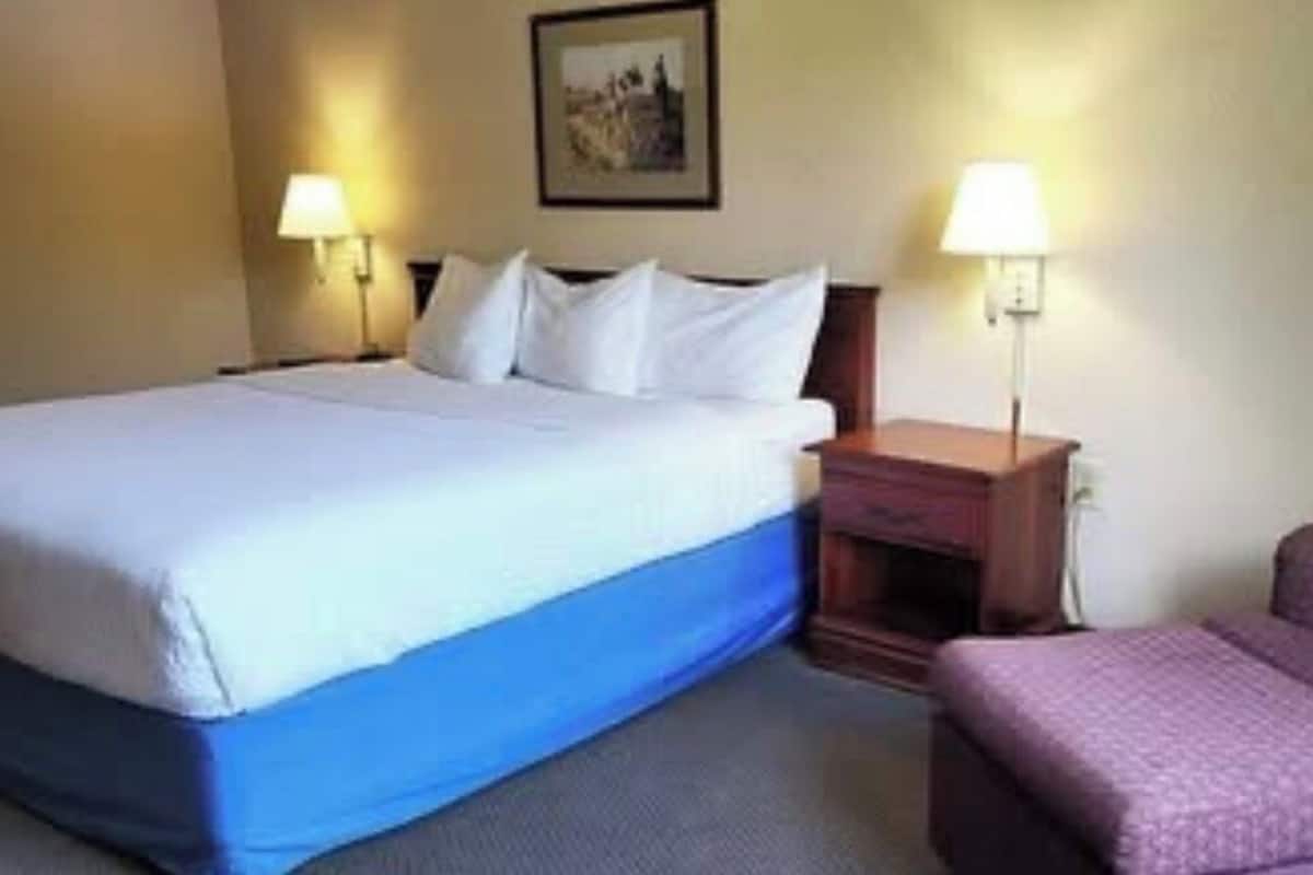 Quarto de hotel com cama, mesas de cabeceira, uma cadeira e um quadro pendurado na parede.