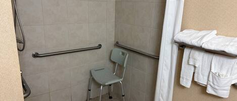 Baño con ducha, barras de apoyo, una silla de ruedas y un perchero para toallas.