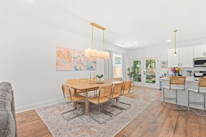 A modern kitchen with white cabinets, a central island, and a dining area with a wooden table and chairs. - New Build | <10 Mins to Folly & <15 to DT (Charleston)