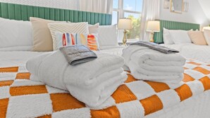 Habitación de hotel con colcha a cuadros anaranjada y blanca, toallas blancas dobladas con cuidado, cabecera tapizada en verde y ventana con vistas a zonas verdes.