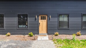 A modern building with a dark exterior, a wooden front door, and small landscaped areas with plants.
