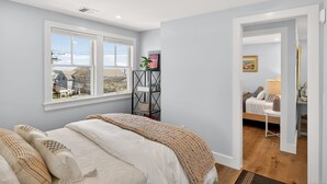 Ein Schlafzimmer mit großem Fenster, einem Bett mit beiger Decke, einem Nachttisch mit Pflanze und Blick auf ein Haus.