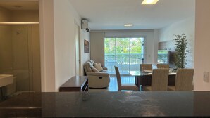 Una sala moderna con un sofá, una mesa de comedor con sillas, una zona de cocina y un balcón con vistas al verdor.
