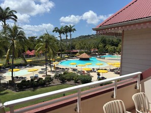 A pool area with sun loungers and umbrellas, surrounded by palm trees and a building with a red roof. - T3 apartment in holiday village - 4/6 persons Sea view & Swimming pool (Rated 3*) (Sainte-Luce)