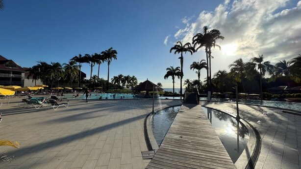 Zona de piscina en un resort con terraza de madera, palmeras y cielo despejado.