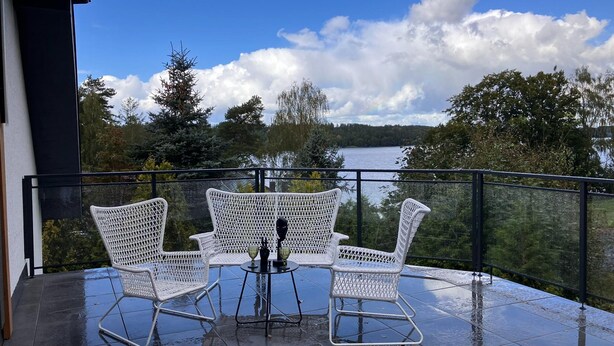 A balcony with two white wicker chairs and a small table, overlooking a lake with trees and a cloudy sky. - Comfortable Apartment With a Private Sauna (gmina Stężyca)