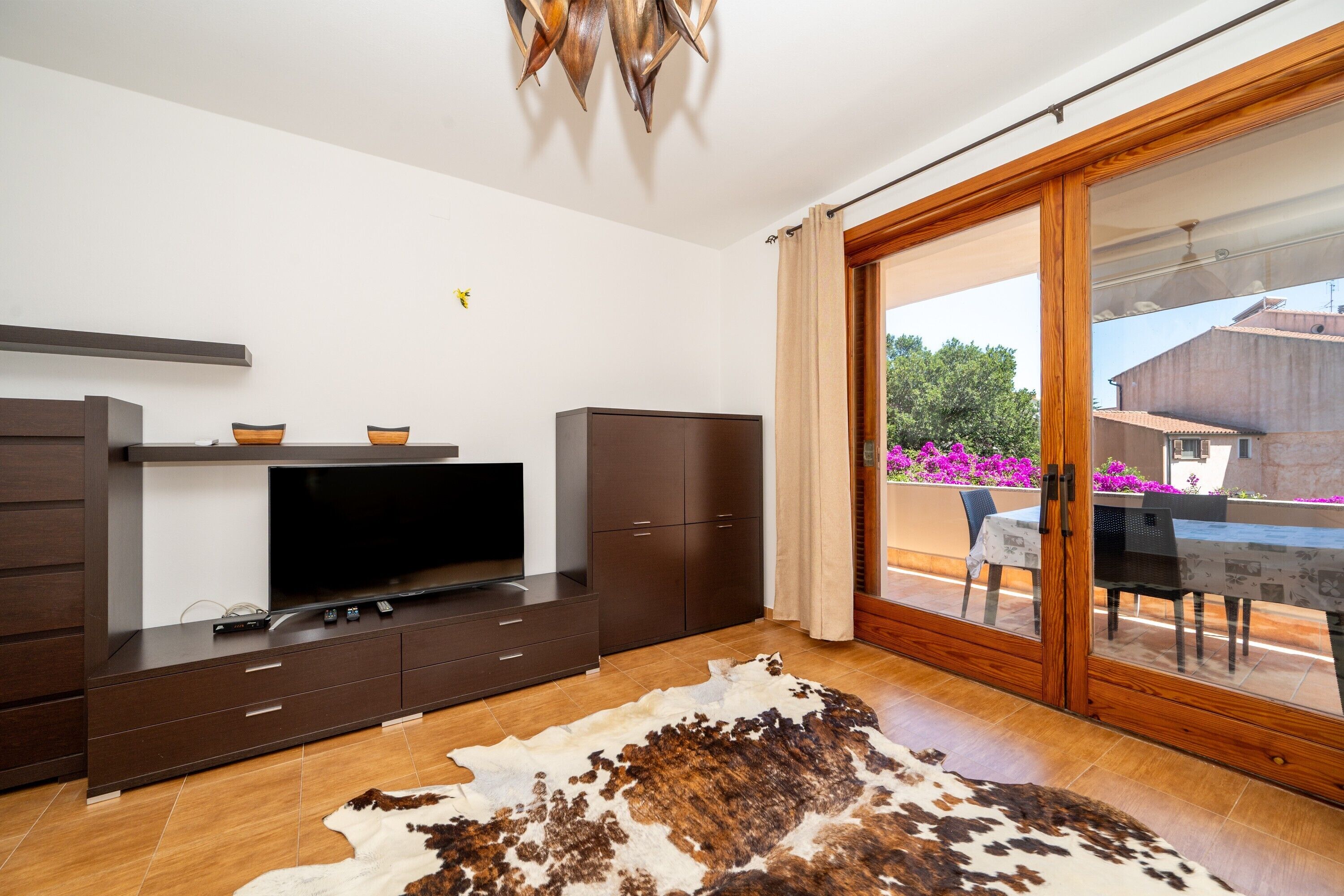 Ein modernes Wohnzimmer mit Flachbildfernseher, dunklen Holzsäschränken, einem Kuhfellteppich und einer Schiebetür zu einer Außenpatio mit Tisch und Stühlen.