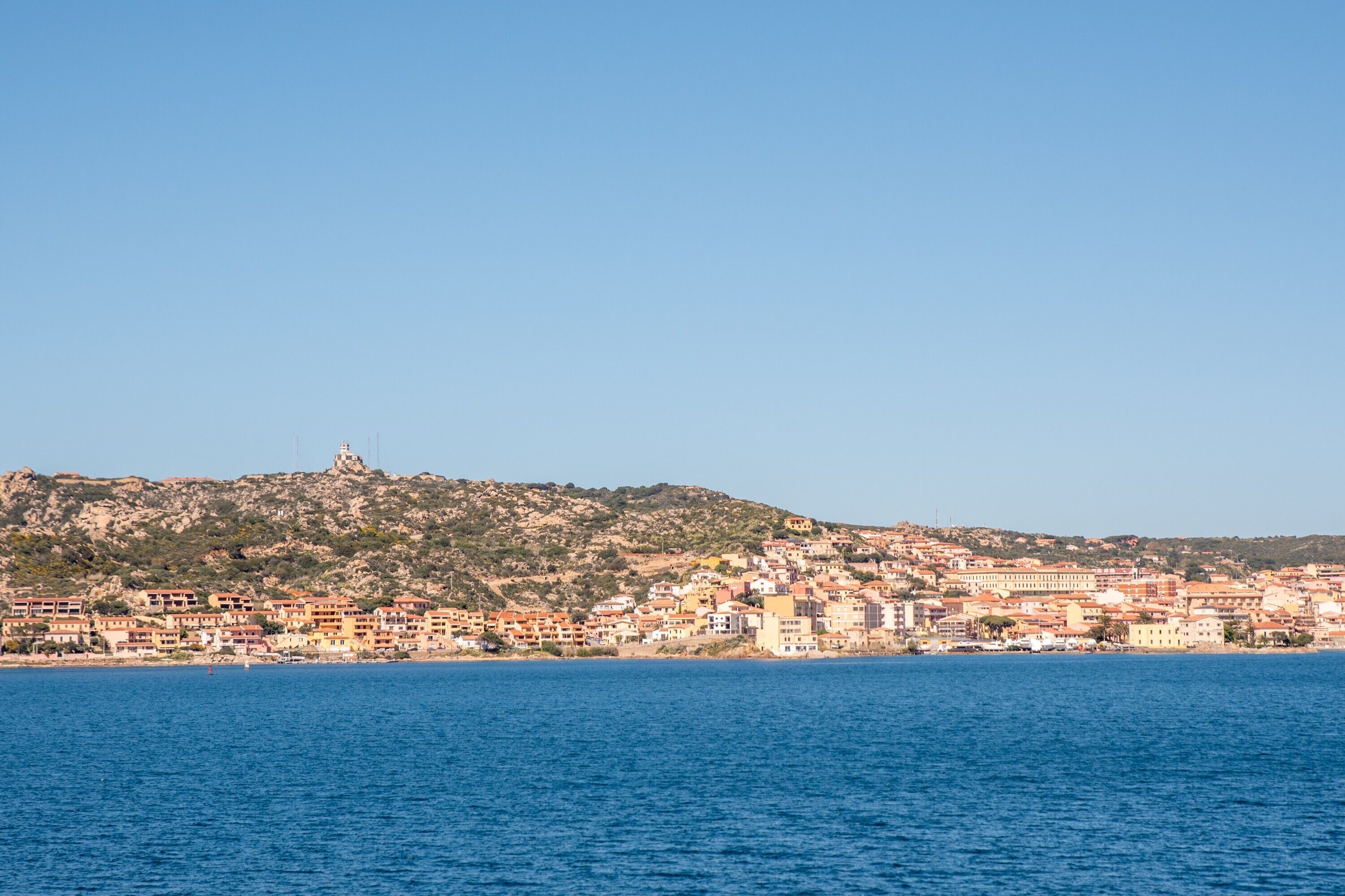 A coastal town with buildings along the waterfront, a hill with a tower, and a clear blue sky.