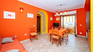 A room with orange walls, a sofa, a dining table with chairs, a TV, and a kitchen area. - Apartment 'Arancio' – Bright 2-Bedroom Retreat in La Maddalena (La Maddalena)