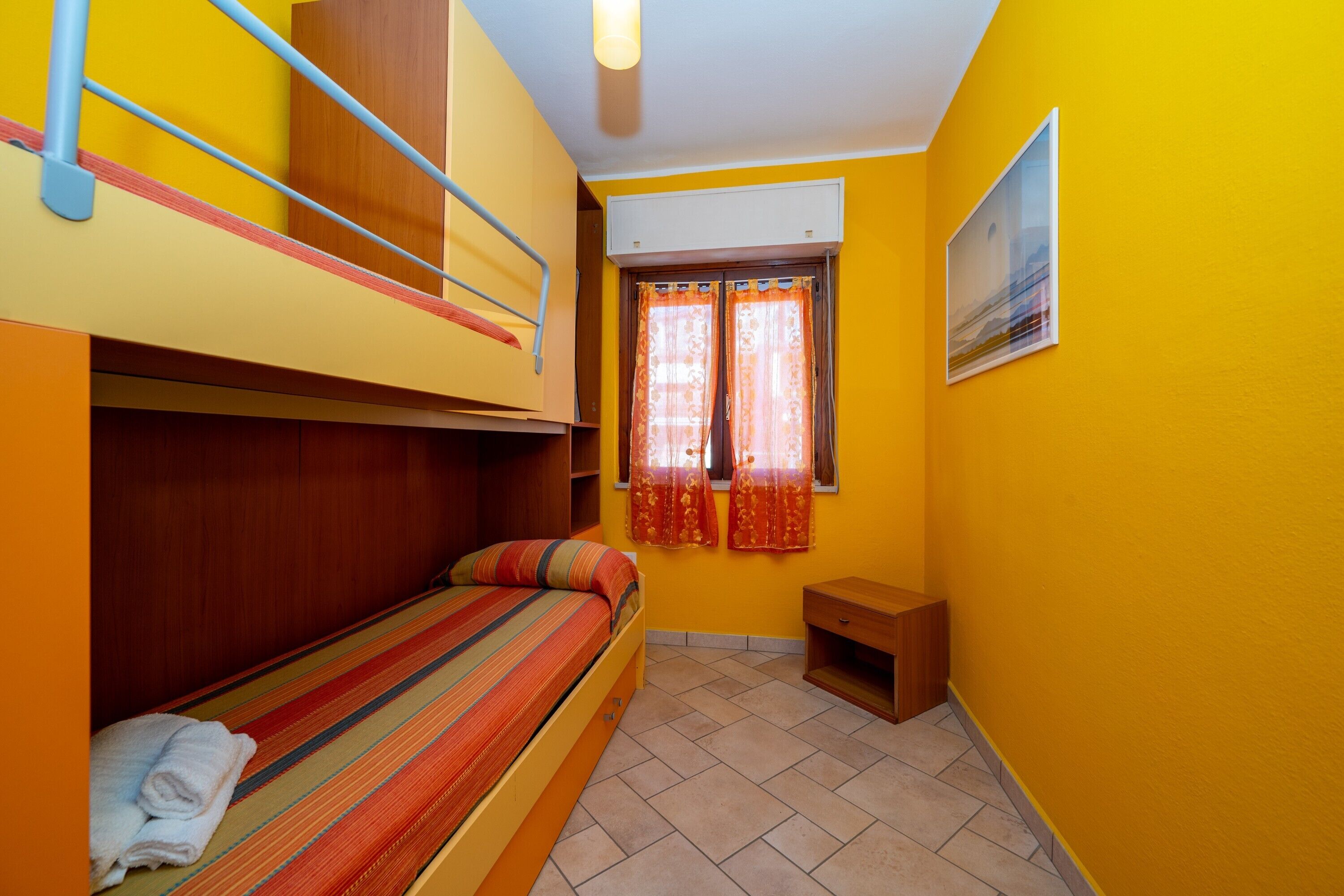 A dormitory room with bunk beds, a window with curtains, a small wooden nightstand, and a ceiling fan.