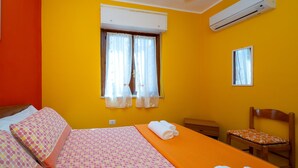 A room with a bed, a chair, a window with curtains, an air conditioning unit, and a small table. - Apartment 'Arancio' – Bright 2-Bedroom Retreat in La Maddalena (La Maddalena)