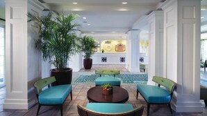Un hall con una recepción, zona de estar con sillas y una mesa, y plantas en macetas grandes.