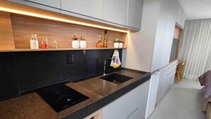 A modern kitchen with a black countertop, two sinks, and a built-in induction cooktop. There are cabinets above the countertop and a small window on the left. - Get One 607 · Flat Corais do Bessa with sea view for 4 people (João Pessoa)