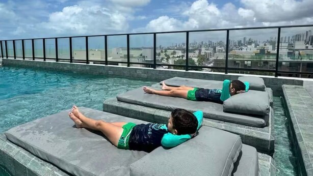Two reclining lounge chairs by a pool with a city skyline view. - Get One 607 · Flat Corais do Bessa with sea view for 4 people (João Pessoa)