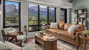 A modern living room with a large window, a brown leather sofa, a wooden coffee table, and a bookshelf. - AlpenPeak Hydeaway at Ascent (Canmore)