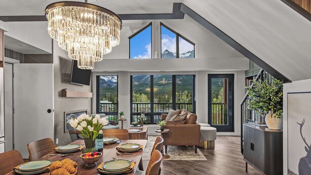 A spacious dining area with a chandelier, a dining table set for a meal, and a view of mountains through large windows. - AlpenPeak Hydeaway at Ascent (Canmore)