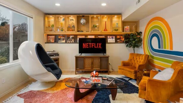 Ein Wohnzimmer mit Glastisch, orangefarbenen Sessel, einem weißen Stuhl, einem Fernseher und einem Bücherregal mit verschiedenen Gegenständen.