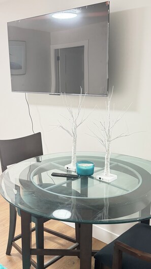 A round glass table with a remote control, a blue candle holder, and two white decorative trees. A flat-screen TV is mounted on the wall above the table. - SERENE COASTAL ESCAPE (Surrey)