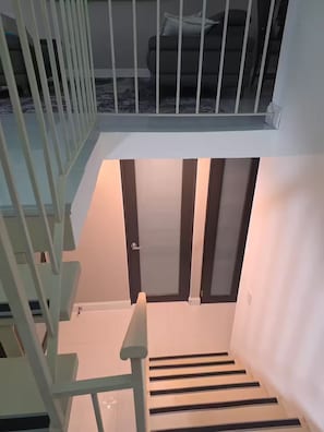 Eine Treppe mit Handlauf, eine Tür am Fußende und Blick auf einen Raum mit Bett und Fenster.
