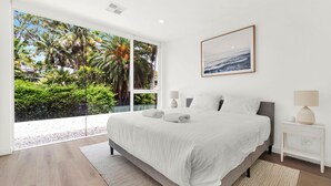 A bedroom with a large bed, a nightstand with a lamp, a framed picture on the wall, and a view of a tropical garden through the window. - Vinny Ocean View by Experience Jervis Bay (Vincentia)