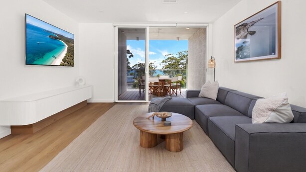 A modern living room with a grey sofa, a wooden coffee table, a TV on the wall, and a sliding glass door leading to an outdoor area. - Vinny Ocean View by Experience Jervis Bay (Vincentia)