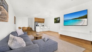 A modern living room with a grey sofa, a dining area, a kitchen, and a large TV displaying a beach scene. - Vinny Ocean View by Experience Jervis Bay (Vincentia)