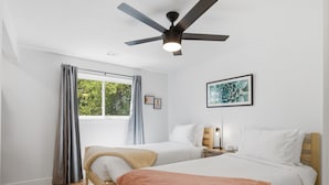 Een slaapkamer met twee bedden, een plafondventilator, een raam met gordijnen, en een lijst met een afbeelding aan de muur.
