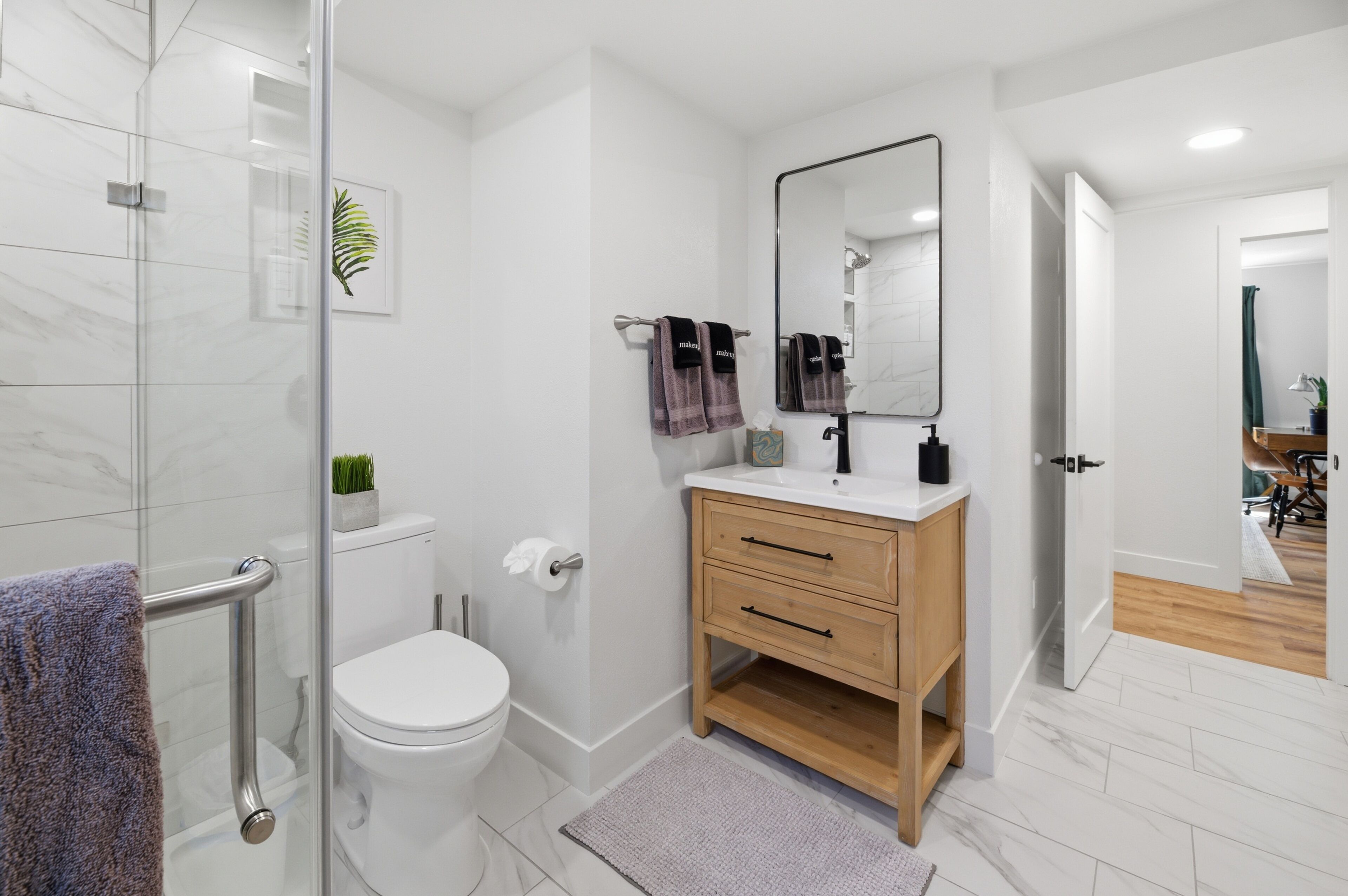 A modern bathroom with a glass shower enclosure, a white toilet, a wooden vanity with a mirror, and a small plant.