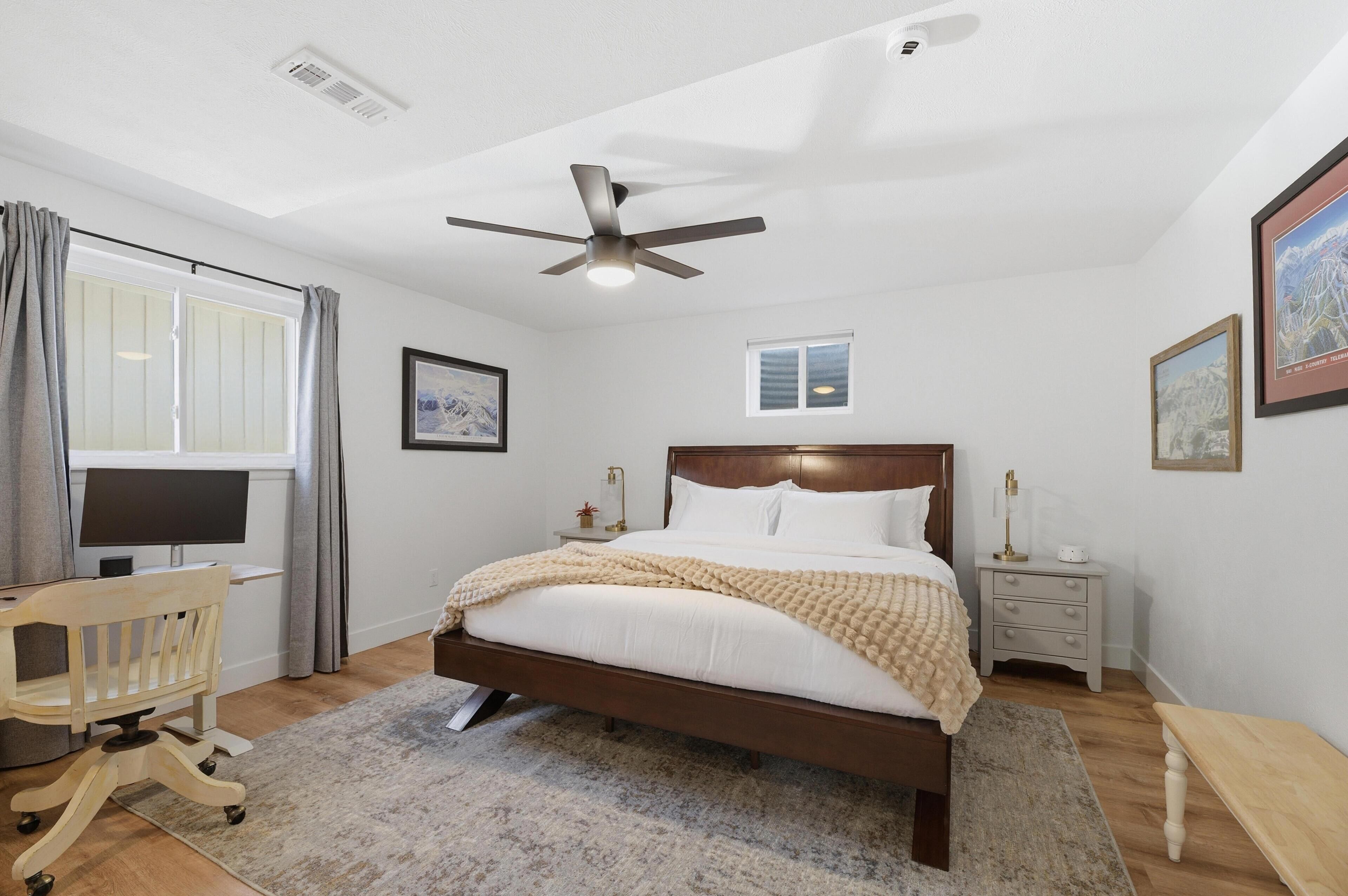 Un dormitorio con cama, escritorio, silla, mesita de noche, ventilador de techo y ventana.